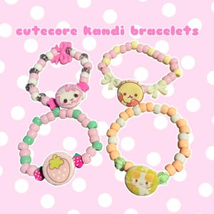 May include: Four colorful kandi bracelets with cute charms and beads. The bracelets feature pink, green, yellow, and orange beads, with charms including a strawberry, a cat, and other kawaii designs. The text "cutecore Kandi bracelets" is at the top.