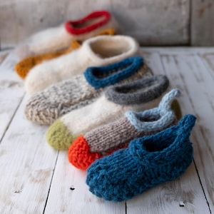 May include: A collection of six hand-knitted slippers arranged in a row. Each slipper features a distinct color combination, including solid blue, orange-toed with brown, light blue, grey, speckled beige with blue trim, and cream with red trim.