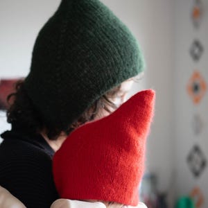 以下が含まれることがあります： 濃いフォレストグリーンのニット製とんがり帽子を被った人物と、鮮やかな赤のニット製とんがり帽子を被った人物。どちらの帽子も、柔らかい質感の糸で編まれており、先端が細くなった特徴的なデザインが施されています。