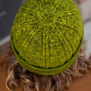 May include: A vibrant olive green knit beanie featuring a complex, textured cable-knit pattern that radiates from the center crown. The hat has a thick, ribbed folded brim and is constructed from a dense, wool-like yarn with visible stitch detail.