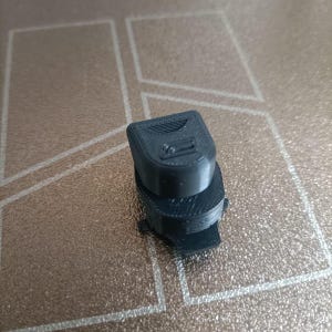Può includere: Un piccolo oggetto nero stampato in 3D, con una parte superiore rettangolare e una base arrotondata. La parte superiore presenta linee parallele e una piccola rientranza rettangolare. Sembra essere un componente di un meccanismo più grande.