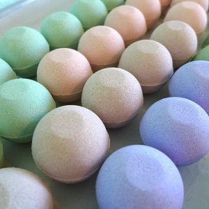 May include: A collection of bath bombs in various shades of green, pink, and purple. The bath bombs are round and smooth, and they are arranged in a row.