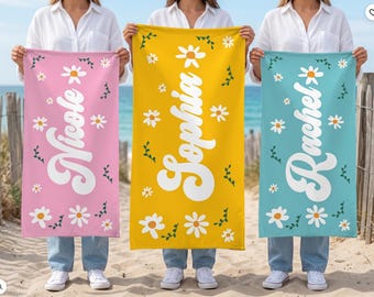 May include: Three personalized beach towels in pink, yellow, and teal, each with a name in white script and daisy accents. The towels are held up by people on a sandy beach.