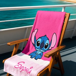 May include: A pink beach towel featuring a blue cartoon character with large ears, resting on a wooden beach chair. The towel has a white stripe and the name "Sophia" in pink script. The chair is on a deck overlooking the ocean.