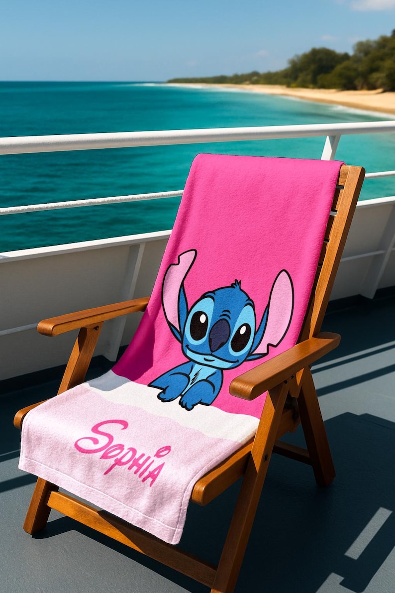 May include: A pink beach towel featuring a blue cartoon character with large ears, resting on a wooden beach chair. The towel has a white stripe and the name "Sophia" in pink script. The chair is on a deck overlooking the ocean.