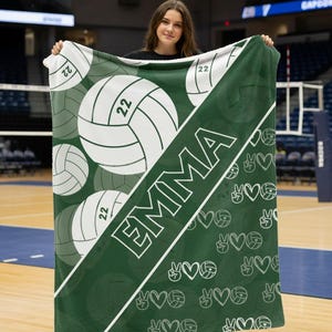 Puede incluir: Una manta de forro polar verde oscuro y blanca con una franja diagonal que muestra el nombre EMMA en letras may&uacute;sculas blancas. El dise&ntilde;o incluye gr&aacute;ficos grandes de voleibol con el n&uacute;mero 22 y un patr&oacute;n repetitivo de corazones y signos de paz.