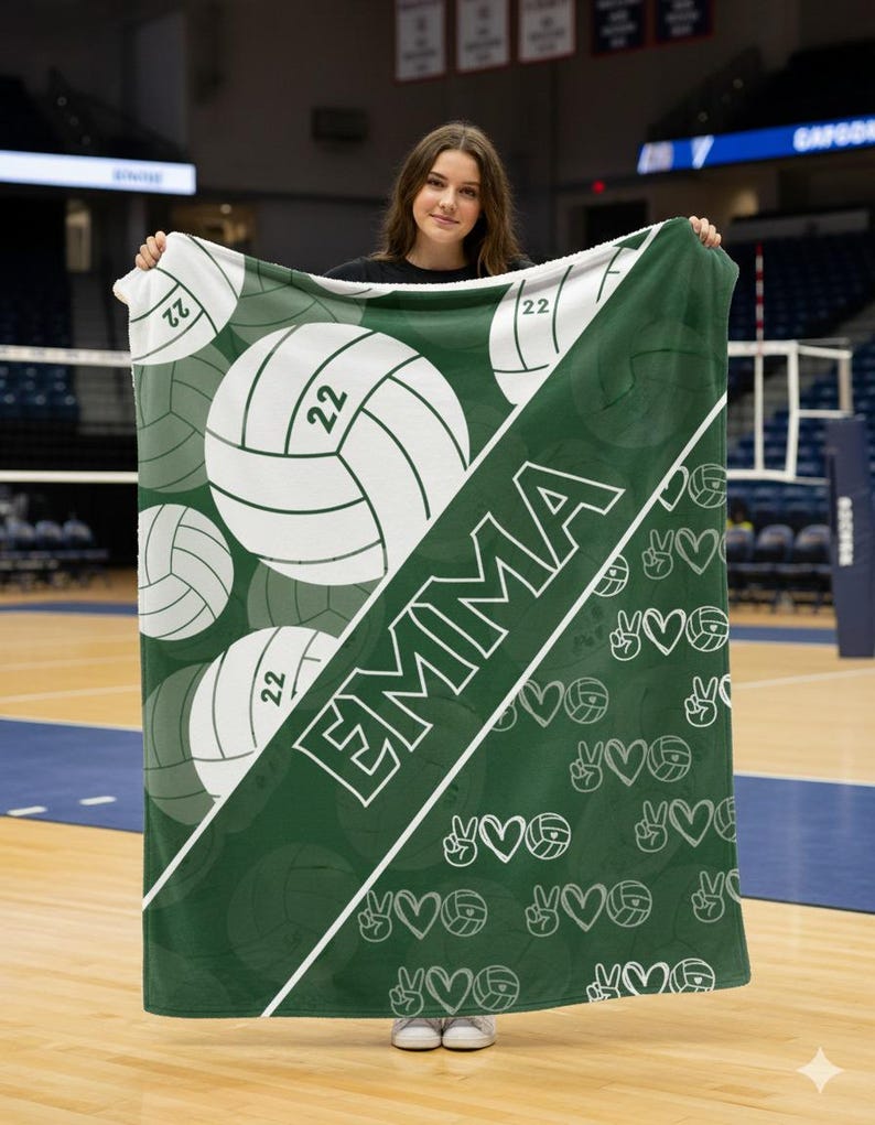 Puede incluir: Una manta de forro polar verde oscuro y blanca con una franja diagonal que muestra el nombre EMMA en letras may&uacute;sculas blancas. El dise&ntilde;o incluye gr&aacute;ficos grandes de voleibol con el n&uacute;mero 22 y un patr&oacute;n repetitivo de corazones y signos de paz.
