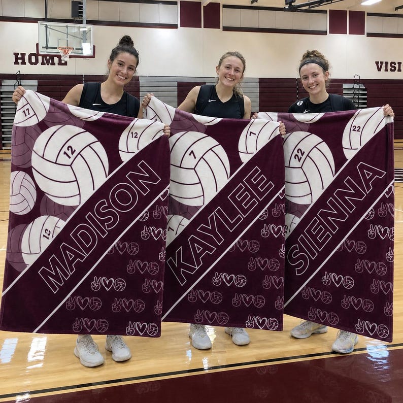 Puede incluir: Tres mantas de forro polar granate y blanco con gr&aacute;ficos de voleibol, los nombres MADISON, KAYLEE y SIENNA, y los n&uacute;meros 12, 7 y 22. Las mantas muestran un patr&oacute;n repetitivo de peque&ntilde;os balones de voleibol, corazones y signos de paz.