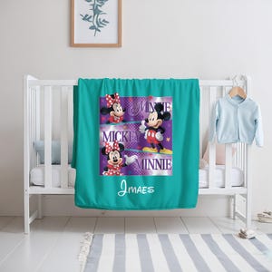May include: A teal blanket featuring Mickey and Minnie Mouse. The blanket has a graphic design with the words "Mickey" and "Minnie" and the name "J MAES". The blanket is draped over a white crib.