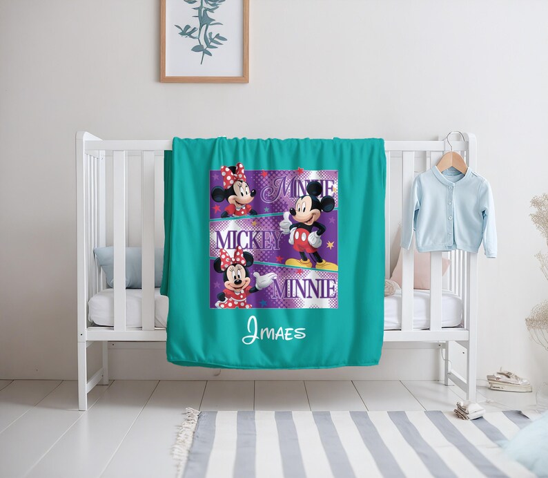 May include: A teal blanket featuring Mickey and Minnie Mouse. The blanket has a graphic design with the words "Mickey" and "Minnie" and the name "J MAES". The blanket is draped over a white crib.