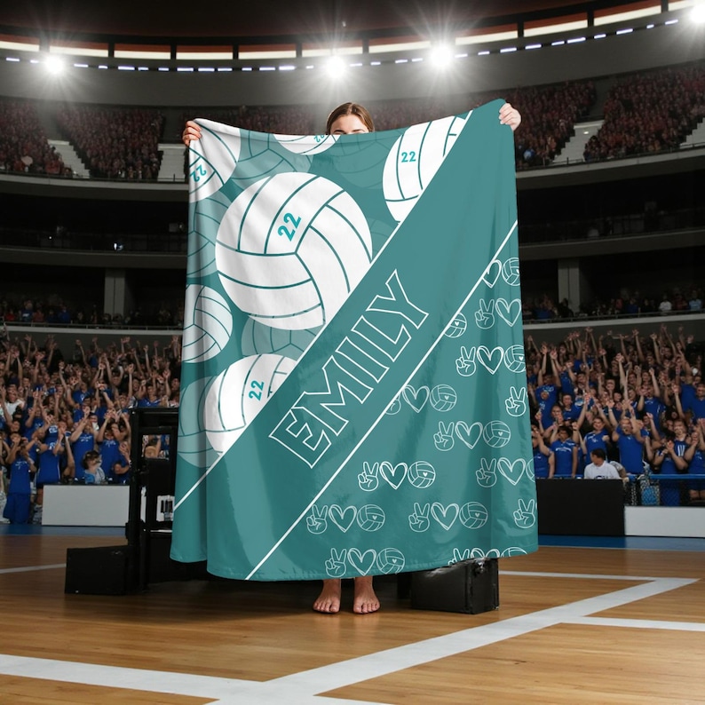 Puede incluir: Una manta de color verde azulado con un patr&oacute;n de balones de voleibol blancos con el n&uacute;mero 22, una franja diagonal blanca con el nombre EMILY en letras may&uacute;sculas blancas y un patr&oacute;n repetido de peque&ntilde;os balones, corazones y signos de paz.