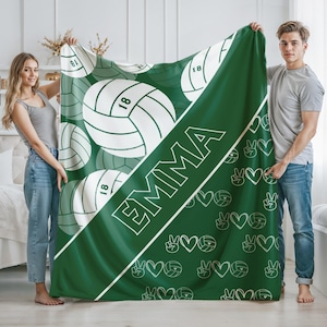 Puede incluir: Una manta de color verde oscuro con una franja diagonal blanca que lleva el nombre EMMA en letras de bloque blancas. El dise&ntilde;o incluye gr&aacute;ficos de voleibol blancos con el n&uacute;mero 18, junto con peque&ntilde;os iconos de corazones y signos de paz.