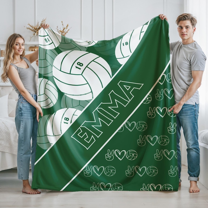 Puede incluir: Una manta de color verde oscuro con una franja diagonal blanca que lleva el nombre EMMA en letras de bloque blancas. El dise&ntilde;o incluye gr&aacute;ficos de voleibol blancos con el n&uacute;mero 18, junto con peque&ntilde;os iconos de corazones y signos de paz.