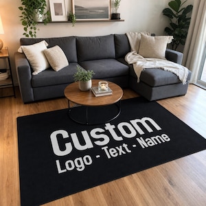 May include: A rectangular black area rug featuring large white text that reads Custom Logo - Text - Name. The rug is positioned on a light-colored wooden floor in front of a dark gray sectional sofa with cream-colored throw pillows and a blanket.