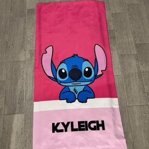 May include: A pink beach towel featuring a blue cartoon character with large ears and big eyes. The character is centered on the towel, above a white stripe and a pink section with the name "KYLEIGH" in black block letters.