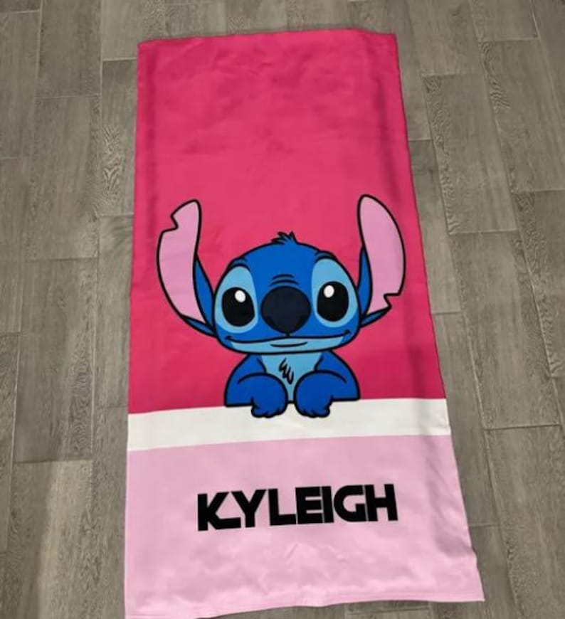 May include: A pink beach towel featuring a blue cartoon character with large ears and big eyes. The character is centered on the towel, above a white stripe and a pink section with the name "KYLEIGH" in black block letters.