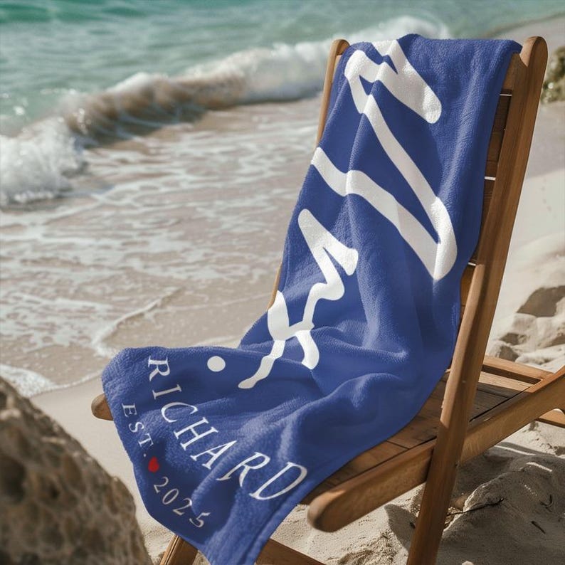 May include: A blue beach towel with white text that says "Richard Est. 2025" and a white abstract design. The towel is folded and sitting on a wooden chair.