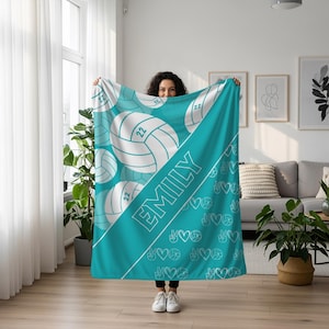 Puede incluir: Una manta de color turquesa con un patr&oacute;n repetitivo de balones de voleibol blancos con el n&uacute;mero 22, una franja diagonal que contiene el nombre EMILY en letras may&uacute;sculas blancas y un patr&oacute;n de signos de paz, corazones e iconos de voleibol.