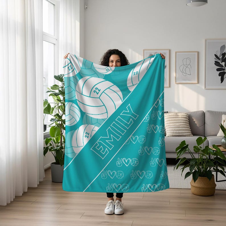 Puede incluir: Una manta de color turquesa con un patr&oacute;n repetitivo de balones de voleibol blancos con el n&uacute;mero 22, una franja diagonal que contiene el nombre EMILY en letras may&uacute;sculas blancas y un patr&oacute;n de signos de paz, corazones e iconos de voleibol.