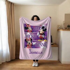 May include: A light purple blanket featuring Mickey and Minnie Mouse. The blanket has a central design with Mickey and Minnie Mouse, the words "Mickey" and "Minnie", and the name "Isabella". The blanket is held up by a person.
