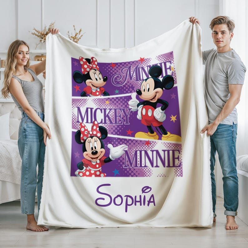 May include: A white fleece blanket featuring Mickey and Minnie Mouse characters in a purple and white design. The blanket has the name "Sophia" in a cursive font. The blanket is held up by two people.