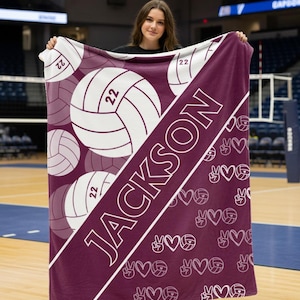 Puede incluir: Una manta rectangular de color granate con una franja diagonal blanca que lleva el nombre JACKSON impreso en letras blancas en negrita. El dise&ntilde;o incluye gr&aacute;ficos de voleibol con el n&uacute;mero 22 y un patr&oacute;n repetitivo de signos de paz y corazones.