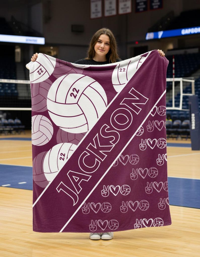 Puede incluir: Una manta rectangular de color granate con una franja diagonal blanca que lleva el nombre JACKSON impreso en letras blancas en negrita. El dise&ntilde;o incluye gr&aacute;ficos de voleibol con el n&uacute;mero 22 y un patr&oacute;n repetitivo de signos de paz y corazones.