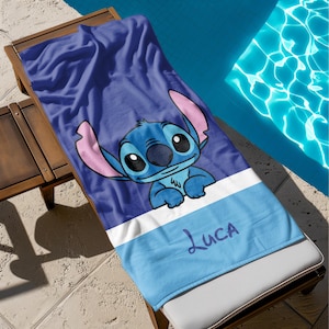 May include: A blue beach towel featuring the character Stitch from Lilo & Stitch. The towel has a large image of Stitch, with pink ears and a blue body, on a dark blue background. The name "Luca" is written in purple script on a light blue stripe.