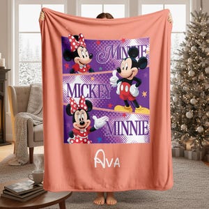 May include: A coral-colored blanket featuring a central design with Mickey and Minnie Mouse characters. The design includes the names "Minnie", "Mickey", and "Minnie" in white text, along with the name "Ava". The blanket has a soft, plush appearance.