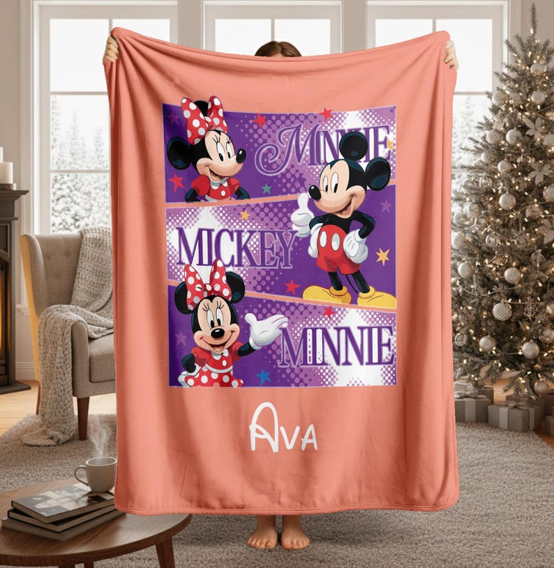 May include: A coral-colored blanket featuring a central design with Mickey and Minnie Mouse characters. The design includes the names "Minnie", "Mickey", and "Minnie" in white text, along with the name "Ava". The blanket has a soft, plush appearance.
