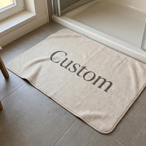 May include: A rectangular beige bath mat with rounded corners featuring the word Custom printed in a dark gray serif font across the center. The mat has a soft, fabric-like texture and is positioned on a gray tiled floor in a bathroom setting.