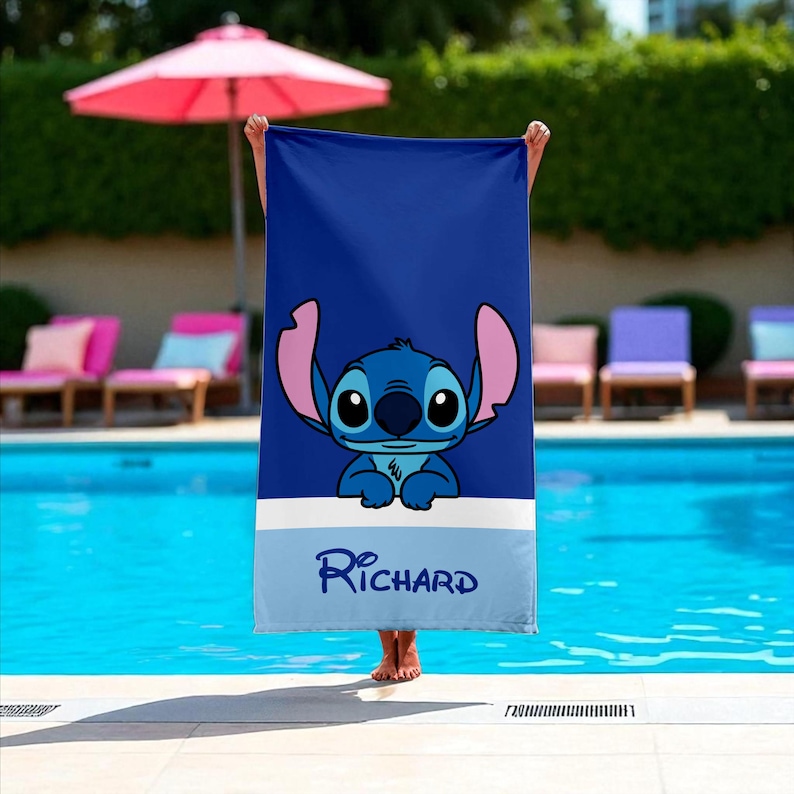 May include: A blue beach towel featuring a cartoon character with large ears and a friendly expression. The towel has a white stripe and the name "Richard" in script. The background includes a swimming pool and lounge chairs.