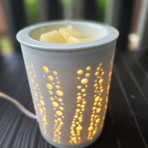 May include: A white ceramic wax warmer with a cylindrical shape and a series of circular cutouts. The warmer is lit from within, and the cutouts emit a warm, yellow glow. The top of the warmer holds several wax cubes.