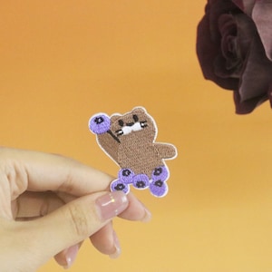 May include: A brown bear embroidered patch with purple blueberries. The bear is holding a blueberry in its right paw and has a happy expression on its face.