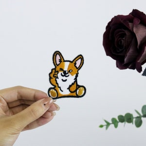 Puede incluir: Parche bordado de un perro corgi marrón y blanco con contorno negro, guiñando un ojo y haciendo un pulgar hacia arriba.