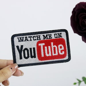 Könnte beinhalten: Ein weißer und schwarzer gestickter Aufnäher mit dem Text "Watch Me On YouTube" in schwarzen Buchstaben. Das YouTube-Logo ist rot mit weißen Buchstaben.