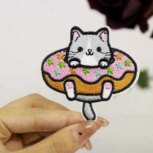 May include: Embroidered iron-on patch featuring a gray cat sitting in a pink donut with colorful sprinkles.
