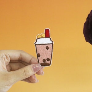 May include: An embroidered patch depicting a boba tea with a red straw and a white lid. The boba tea is filled with brown tapioca pearls.