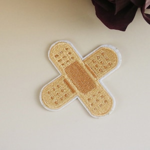 May include: A beige fabric patch shaped like a bandage with white stitching around the edges. The patch is embroidered with a brown design that resembles a bandage.