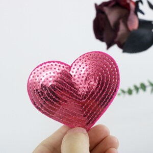 May include: A pink heart-shaped sequin patch. The sequins are arranged in a pattern that creates a heart shape.