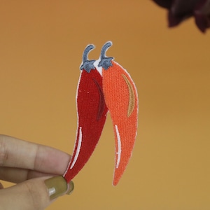Könnte beinhalten: Zwei gestickte Chilischoten, eine rote und eine orange, mit weißen Nähten. Die Chilischoten sind oben zusammengenäht.