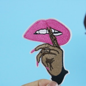 May include: Pink embroidered lips with a finger over them, saying "Shhh" in brown embroidery. The finger is part of a brown hand with black fingernails.