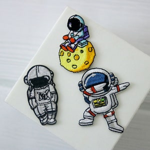 Könnte beinhalten: Drei gestickte Bügelbilder mit Astronauten in verschiedenen Posen. Ein Astronaut sitzt auf einem gelben Mond, ein anderer steht mit ausgestreckten Armen da und der dritte steht mit den Armen an den Seiten.