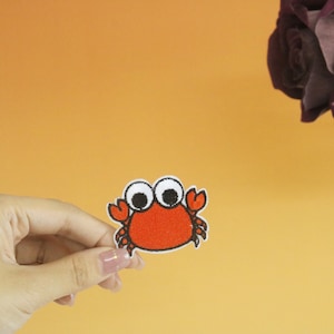Op de afbeelding: Een rode en witte cartoon-krab met grote ogen. De krab is geborduurd op een stoffen patch.