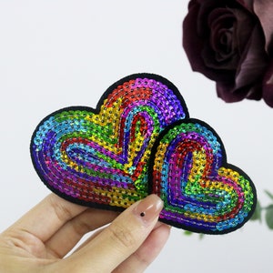 May include: Two heart-shaped patches with rainbow-colored sequins and black borders. The sequins are arranged in concentric patterns, creating a shimmering effect. The patches are held against a white background.