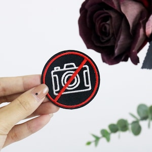Może przedstawiać: Naszywka haftowana w kolorze czarnym i czerwonym z białym aparatem fotograficznym w czerwonym kółku z czerwoną linią przechodzącą przez nią. Naszywka jest trzymana przez rękę.