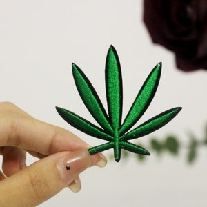 Könnte beinhalten: Grüner gestickter Marihuana-Blatt-Patch. Der Patch wird in einer Hand gehalten.
