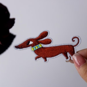 Puede incluir: Parche bordado de un perro salchicha en tonos marrones con borde blanco. El perro tiene orejas largas, un collar azul con detalles amarillos y una cola rizada. El parche se sostiene sobre un fondo blanco.