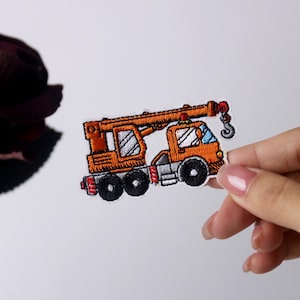 May include: An orange embroidered iron-on patch of a crane truck with a white cab and black wheels.