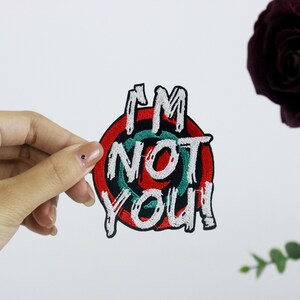 May include: Embroidered patch with the text "I'M NOT YOU!" in white, black, red, and green. The patch has a circular design with a target-like appearance. The patch is held by a hand against a white background.